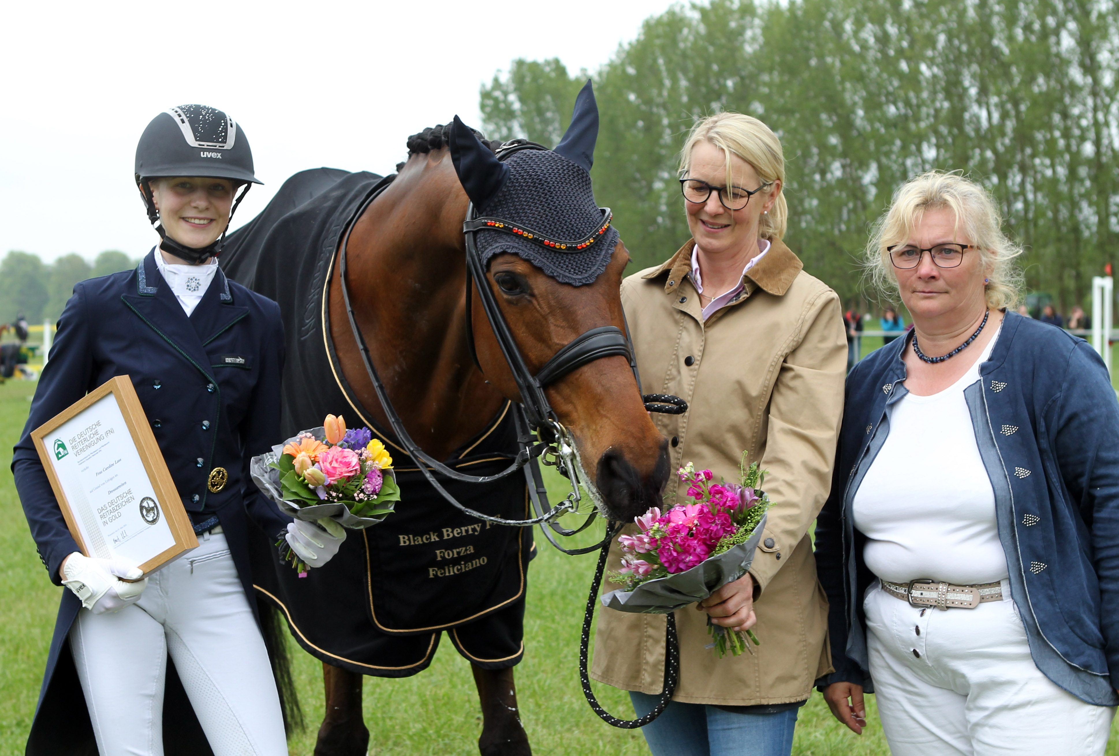 Die 21-jährige Caroline Lass aus Tempel, die für die RSG Wöpkendorf reitet, bekam das Goldene Reitabzeichen. Turnierleiterin Birca Roos (r.) gratuliert und Mutter Inga Lass lässt Feliciano am Blumenstrauß schnuppern. © Jutta Wego